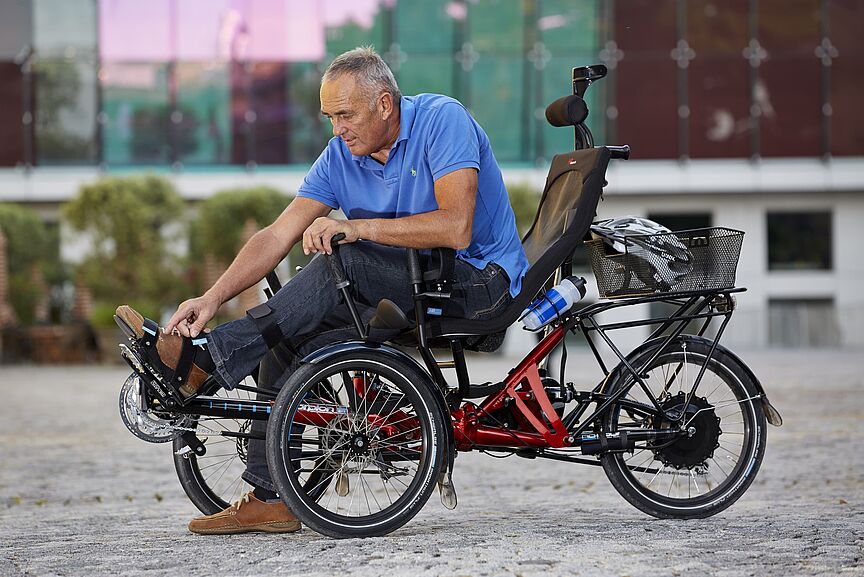 Spezialrad für Menschen mit besonderen Bedürfnissen Ein Mann in blauem Poloshirt sitzt auf einem roten dreirädrigen Elektrofahrrad mit schwarzer Rückenlehne und Korb. Das Spezialrad steht auf einem gepflasterten Platz vor einem modernen Gebäude mit grünen Fenstern.