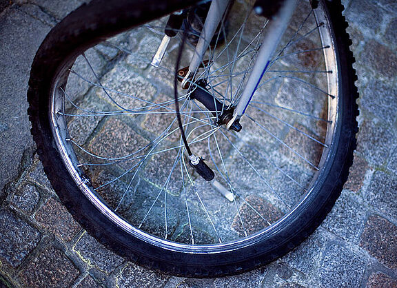 Nahaufnahme eines Fahrradvorderrads auf Kopfsteinpflaster. Die Felge ist verbogen und mehrere Speichen sind gebrochen oder hängen lose herab. Das Rad steht schief auf dem grauen Pflaster und ist offensichtlich nach einem Unfall nicht mehr fahrbereit.