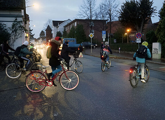 Mehrere Jugendliche fahren auf dem Rad über eine Straße, während es morgens noch dämmert. Sie tragen Winterjacken und Rucksäcke, einige schützen ihren Kopf mit einem Helm. Die Lampen an den Rädern leuchten. Im Hintergrund warten Autos mit eingeschalteten Scheinwerfern an der Straße. Straßenlaternen erhellen die Szene.